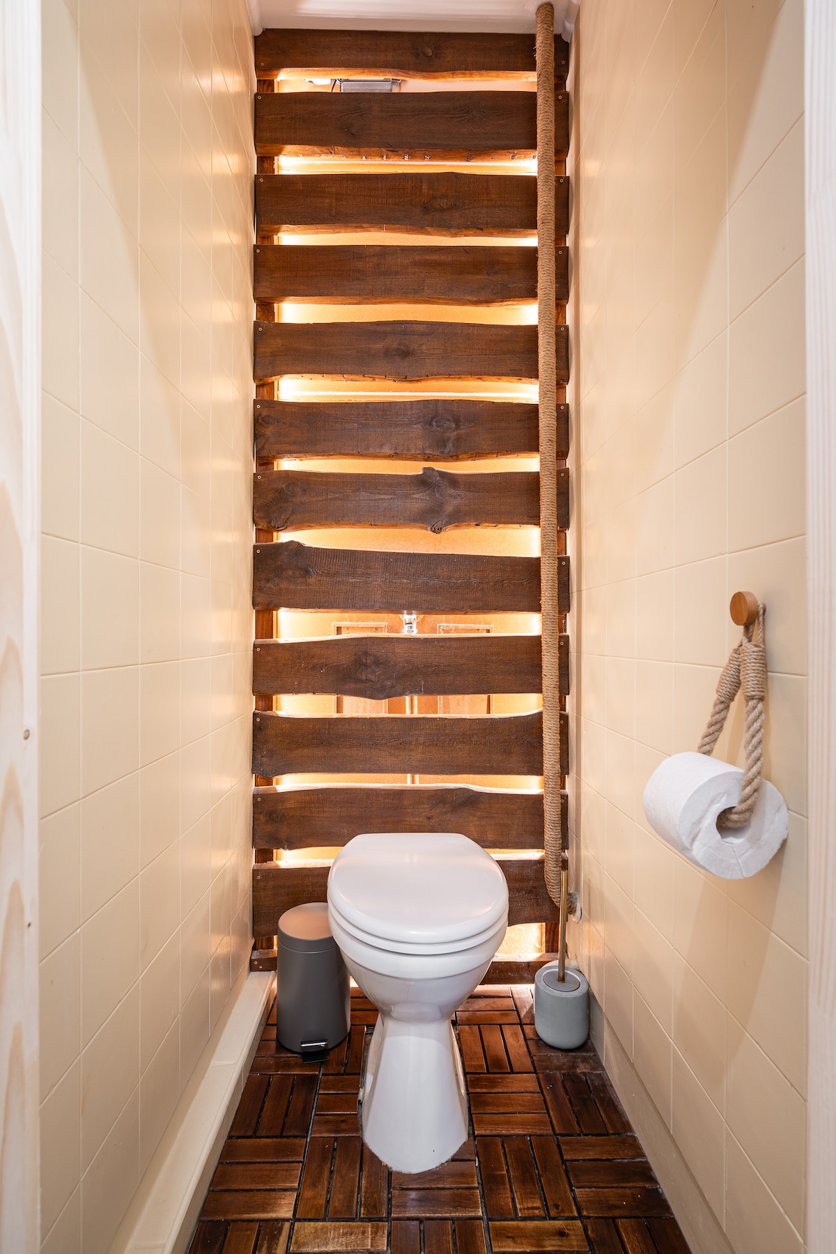 The bathroom features a modern toilet set against a backdrop of wooden slats allowing natural light to filter through. The floor consists of brown tiles, adding warmth to the space. Essential items, such as toilet paper and a small waste bin, are also present.