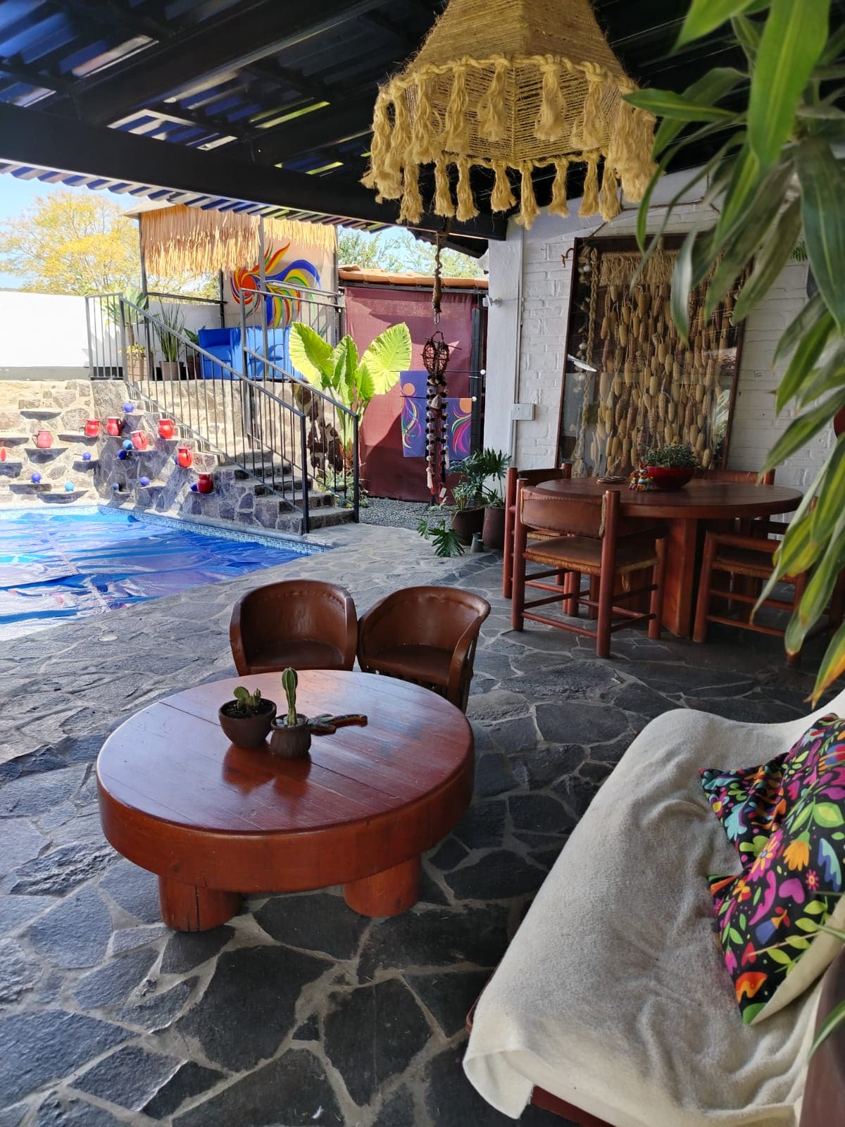 An outdoor seating area features a wooden table surrounded by chairs, with a colorful tapestry in the background. A cozy sofa adorned with a patterned cushion rests on stone flooring, offering a view of the private pool and lush greenery.