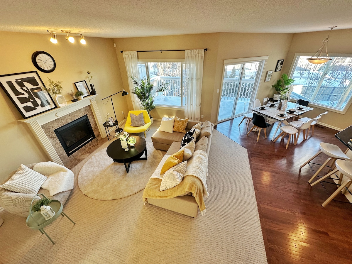 An inviting living space features a light-colored sofa and armchair arranged around a circular area rug. A fireplace is positioned beside a large window offering natural light. A dining area with a round table and chairs is visible, complemented by nearby greenery and decorative elements.