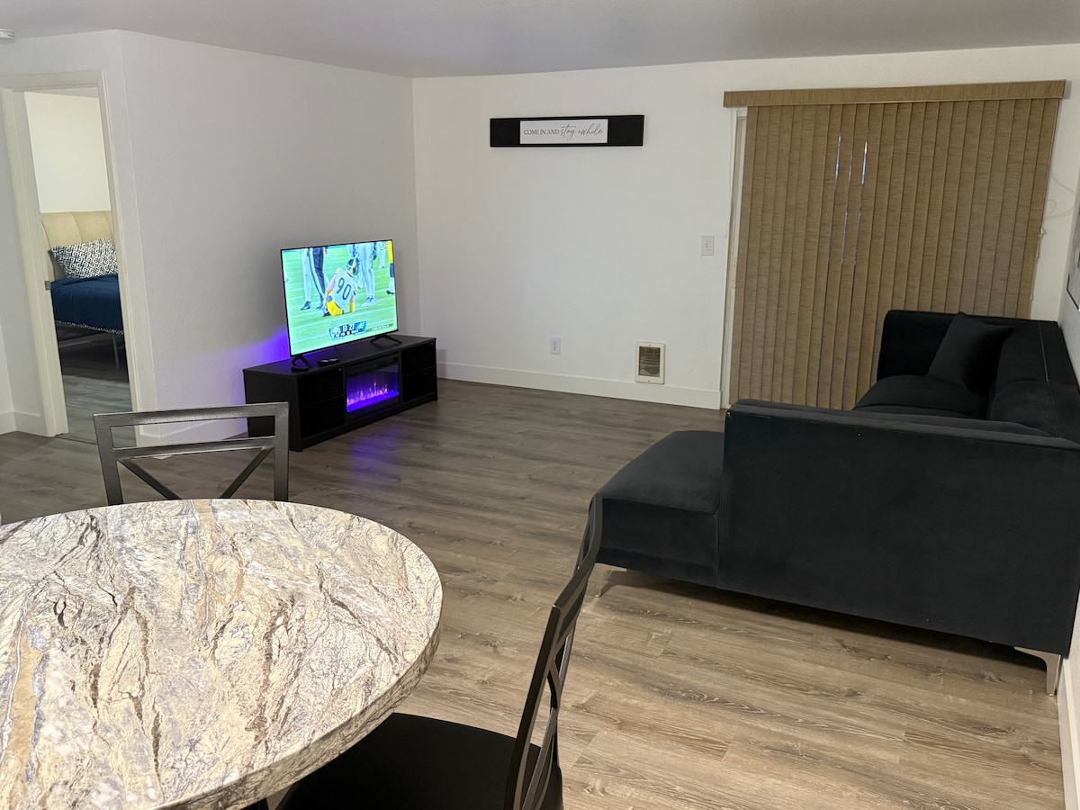 An open living area features a dark sofa facing a TV mounted above a low console unit. Natural light enters through a partially closed window with horizontal blinds. A round dining table with a marbled surface and black chairs is positioned nearby, accentuating the modern decor.