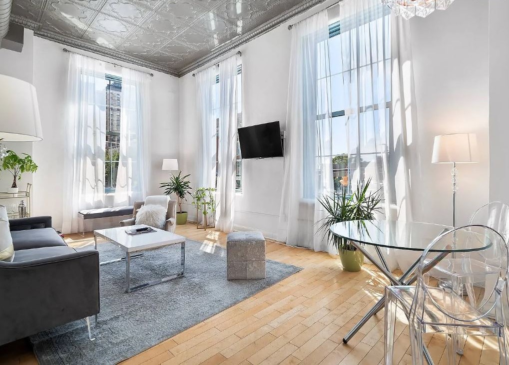 A bright living area features large windows allowing natural light to fill the space. A cozy seating arrangement includes a gray sofa and a glass coffee table on a soft area rug. Decorative plants and contemporary lighting enhance the ambiance, alongside a modern TV mounted on the wall.