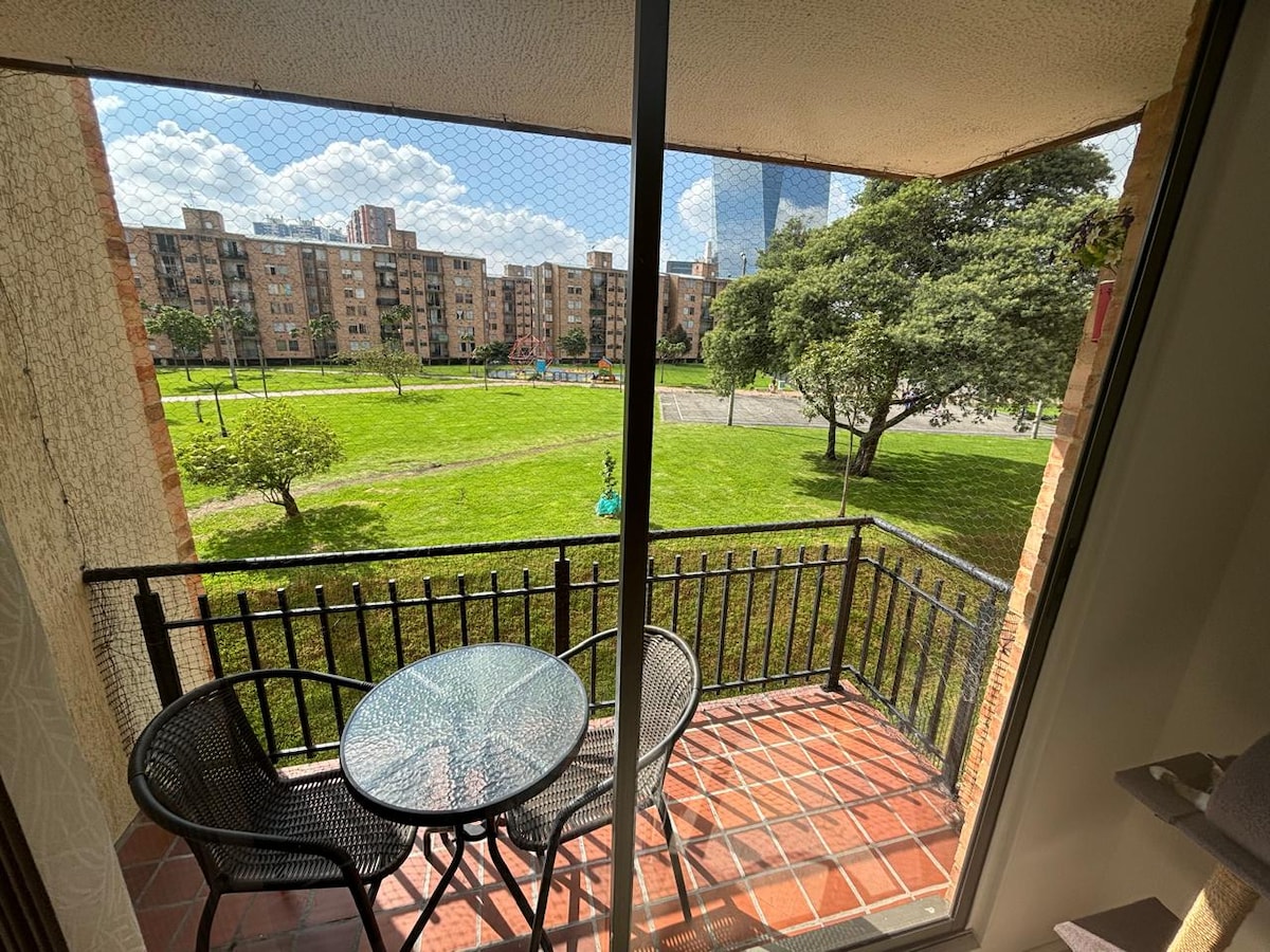 A balcony with a glass table and two black chairs overlooks a green area surrounded by residential buildings. The ground below features well-maintained grassy patches and a few trees, creating a serene outdoor setting. Bright sunlight illuminates the space, enhancing the view.