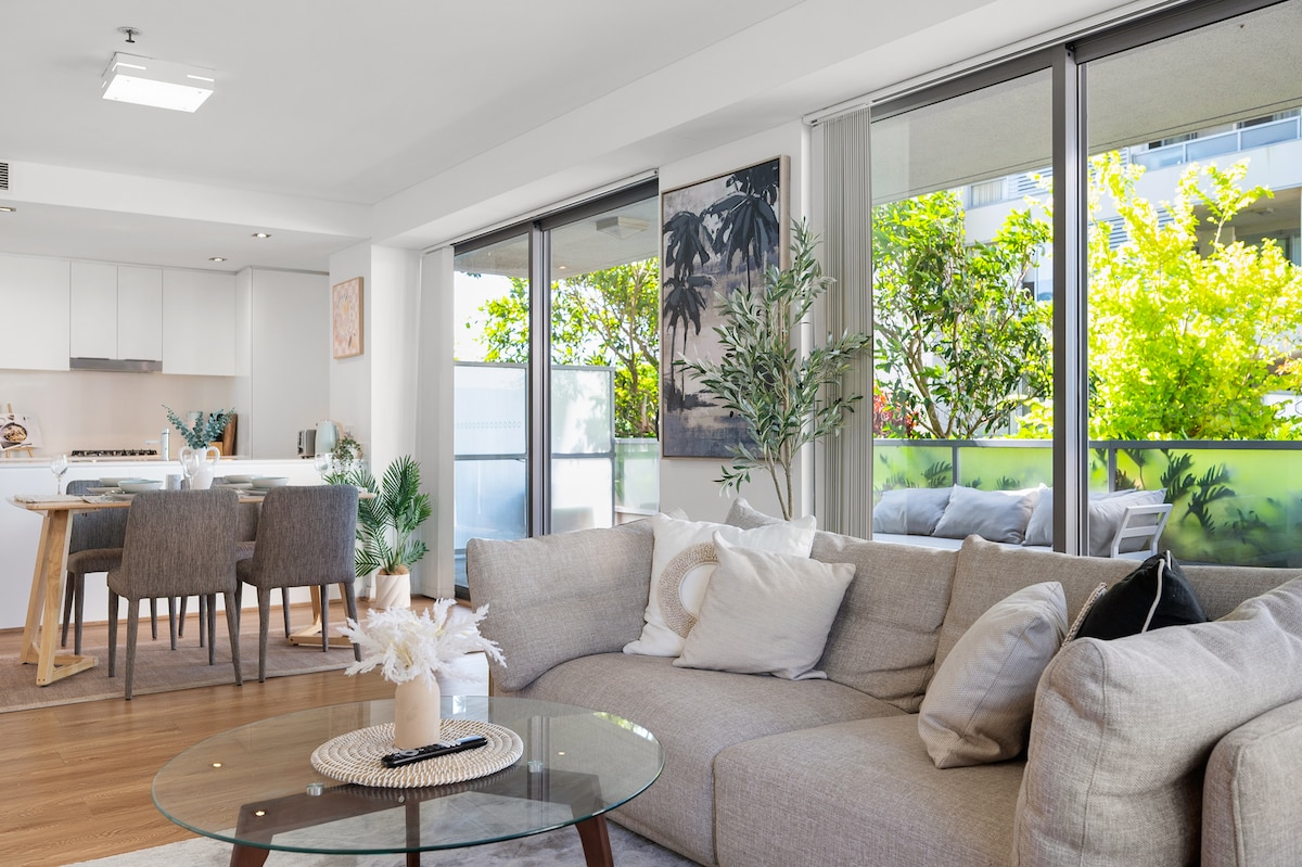 A bright living area is featured with large glass sliding doors that open to a balcony, allowing natural light to enhance the space. A comfortable sofa with assorted cushions is paired with a glass coffee table. Indoor plants and tasteful wall art complete the inviting ambiance.