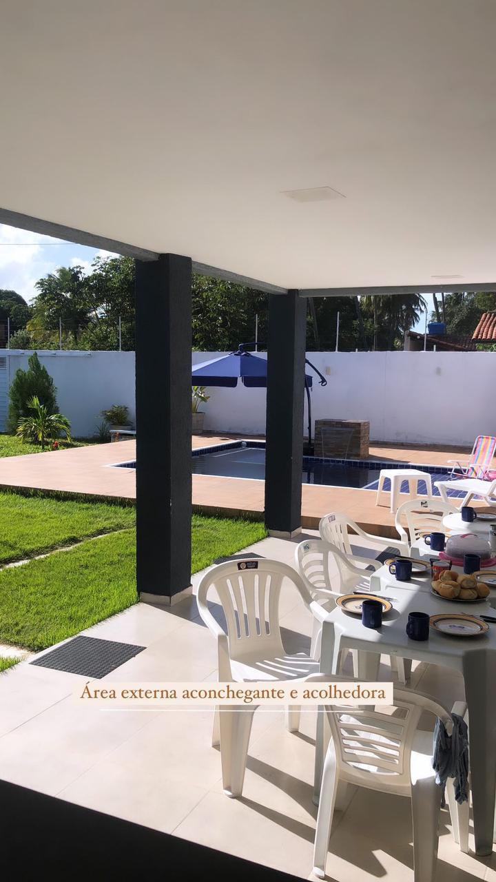 An outdoor area features a spacious patio with white chairs arranged around a table set for dining. A pool is visible in the distance, surrounded by greenery and a partially shaded lounging area. Sunlight enhances the tranquil setting.