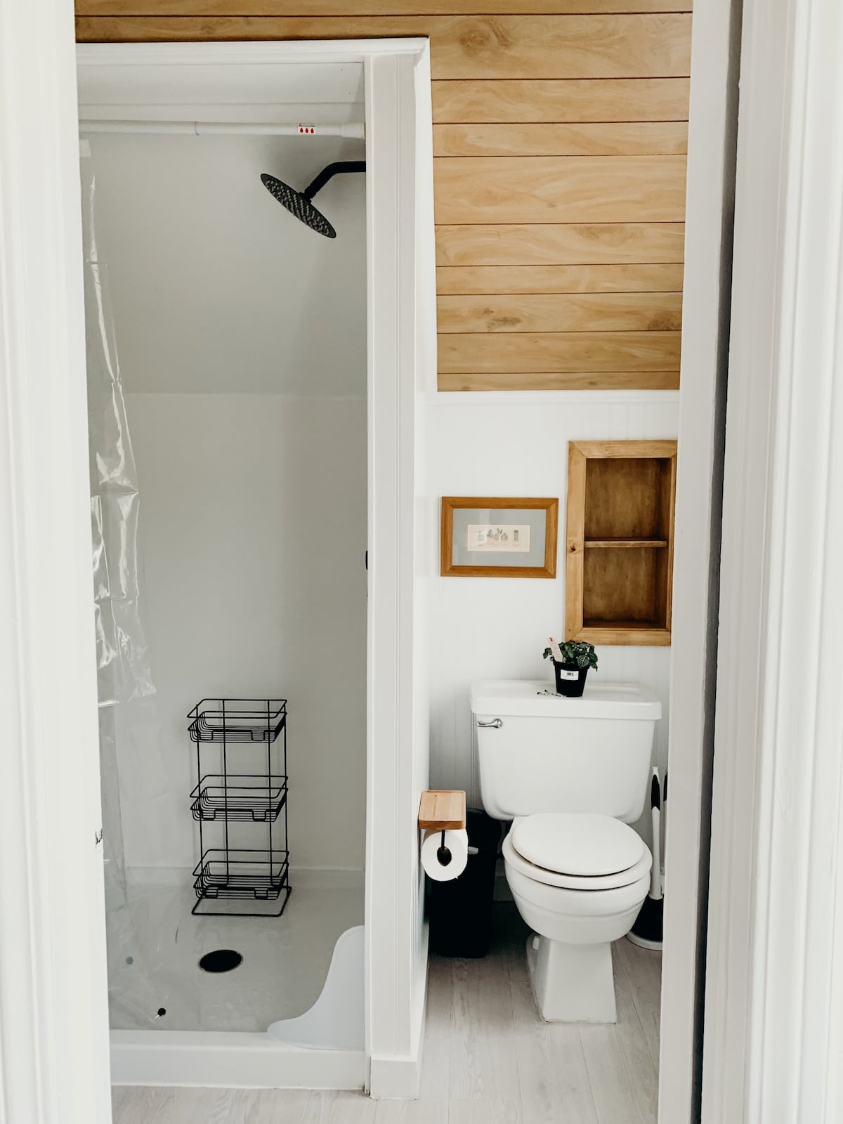 A light and compact bathroom is visible, featuring a walk-in shower equipped with an overhead rain showerhead. A modern toilet is placed adjacent to the shower, alongside a wooden cabinet with shelves. Natural light illuminates the space, highlighting the neutral color palette and warm wooden accents.