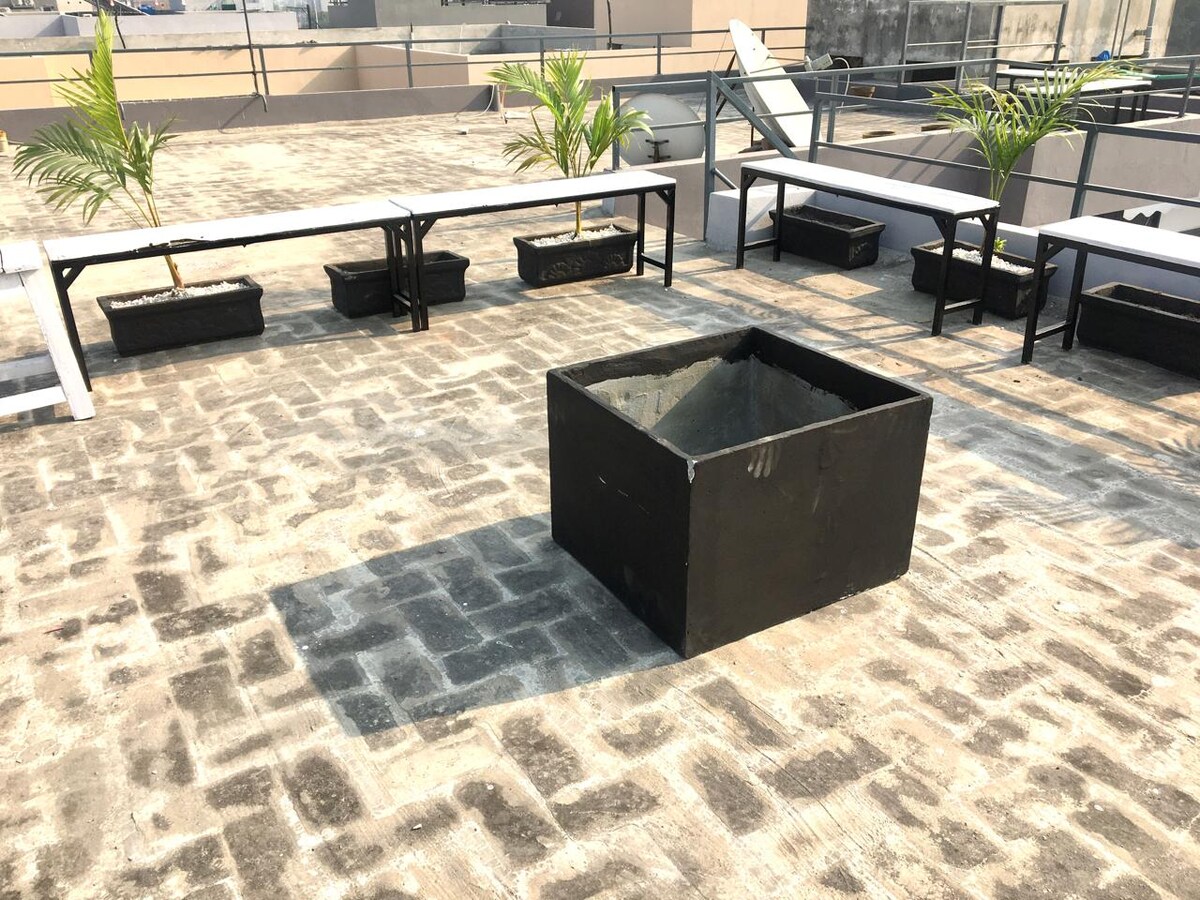 The rooftop patio features a central fire pit with surrounding seating arrangements. Potted plants add greenery to the space, while long benches provide comfortable spots for guests. The textured flooring enhances the outdoor ambiance, creating a setting for small gatherings.