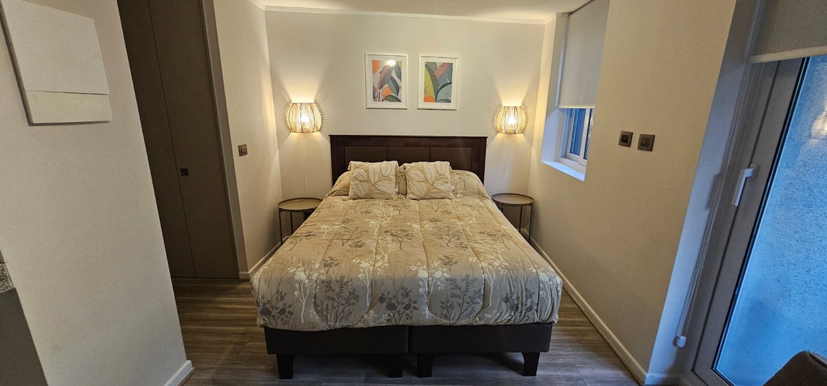 A spacious sleeping area features a large bed with a decorative floral quilt. Two bedside tables hold stylish lamps, while framed artwork adorns the walls. Natural light enters through a window, complemented by light-colored shades.