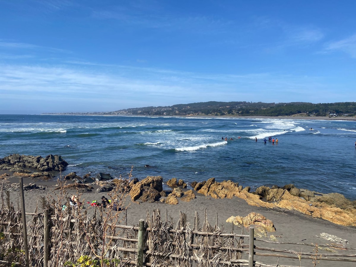 A serene beach scene features gentle waves lapping against rocky outcrops. Groups of people can be seen enjoying the water, while distant hills create a tranquil backdrop under a clear blue sky. The sandy shore is interspersed with patches of vegetation and wooden fencing.
