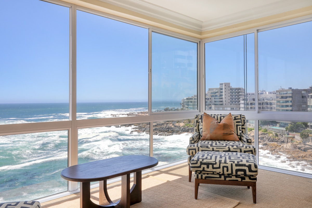 A sunlit seating area features a patterned armchair positioned beside a glass wall, offering expansive views of the ocean waves and coastal landscape. A wooden coffee table complements the space, while natural light fills the room, enhancing the sense of openness.
