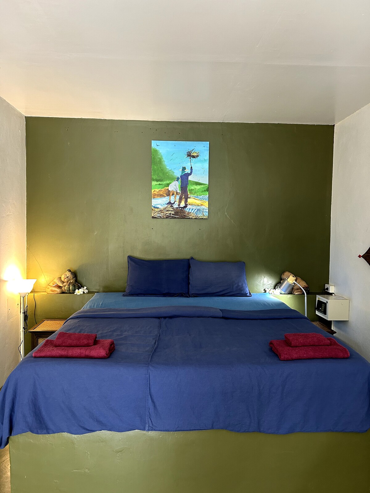 The sleeping area features a spacious bed with blue bedding and accent pillows. A vibrant painting hangs above the bed, complemented by warm lighting from bedside lamps. Two neatly folded towels are placed at the foot of the bed, enhancing the cozy appearance of the room.