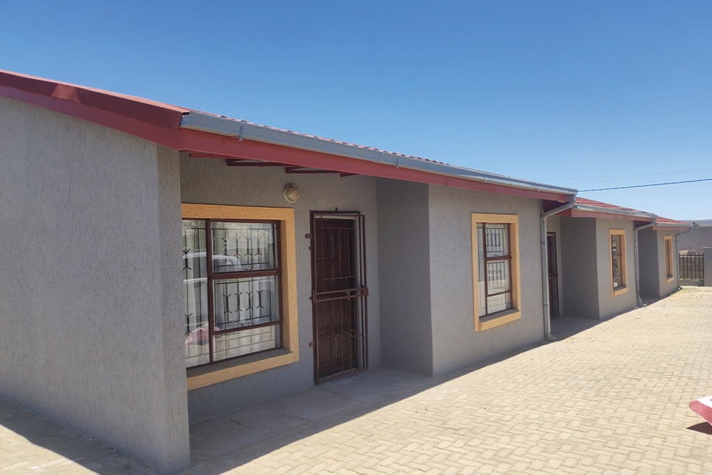 The exterior of a single-story house is showcased, featuring a grey facade with wooden trim around the windows. The red roof provides a contrasting accent. The paved pathway leads to a prominent front door, while three evenly spaced windows offer natural light.