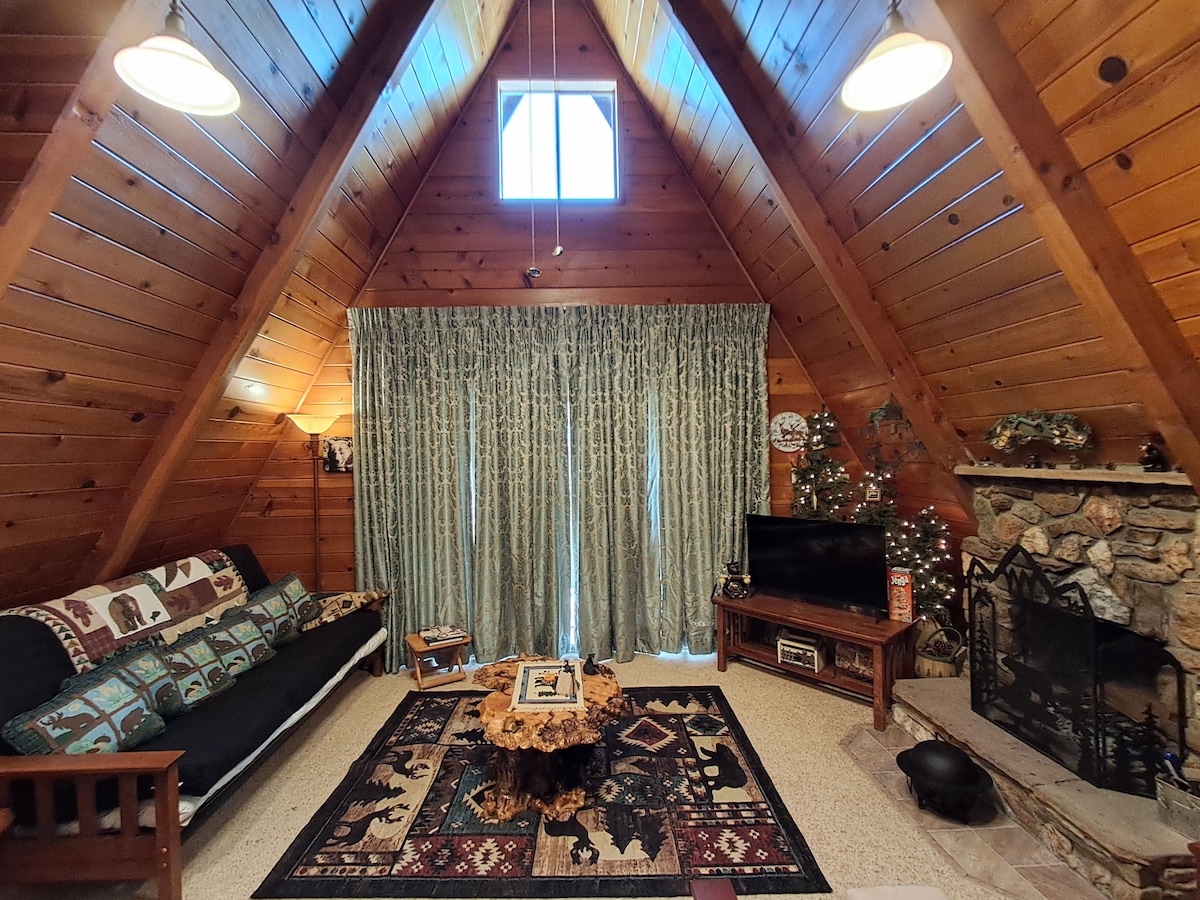 A cozy living area is highlighted by a high, wood-paneled ceiling and a stone fireplace. A dark sofa is adorned with bear-themed cushions, complemented by a large area rug. Natural light filters through a small window above, while elegant curtains frame the space.