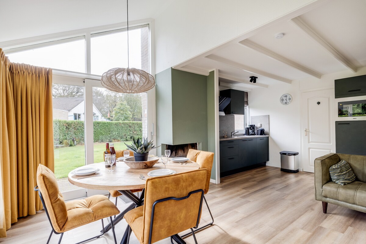 A modern dining area features a round wooden table surrounded by four upholstered chairs. Natural light floods the space through large glass doors that lead to a private garden. The adjacent kitchen is equipped with sleek cabinetry and modern appliances, contributing to a bright and open design.