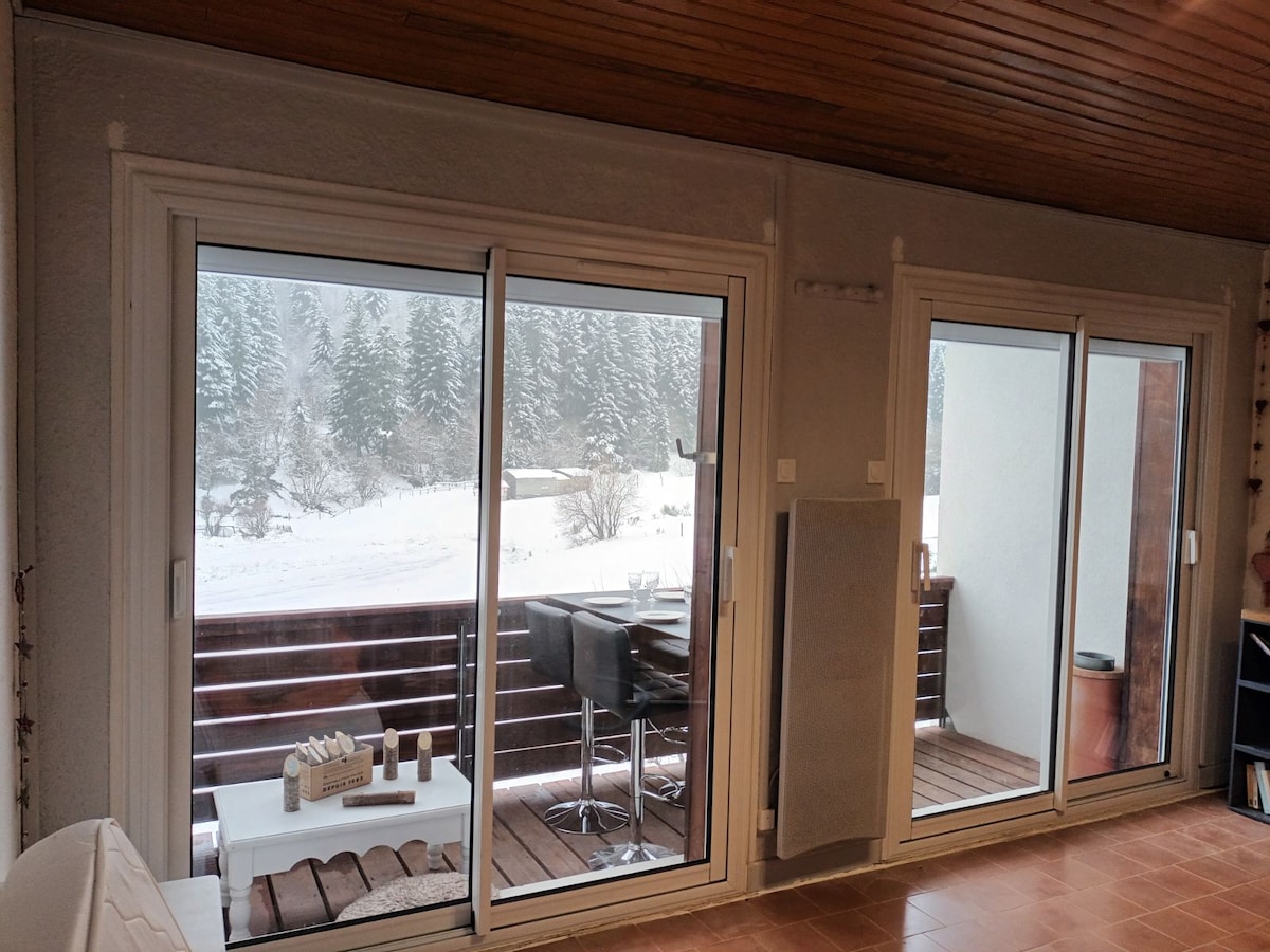 A spacious living area is highlighted by large sliding glass doors that open to a balcony. Snow-covered trees are visible outside, while a small outdoor table and chairs are positioned on the balcony, creating a connection to the serene winter landscape.