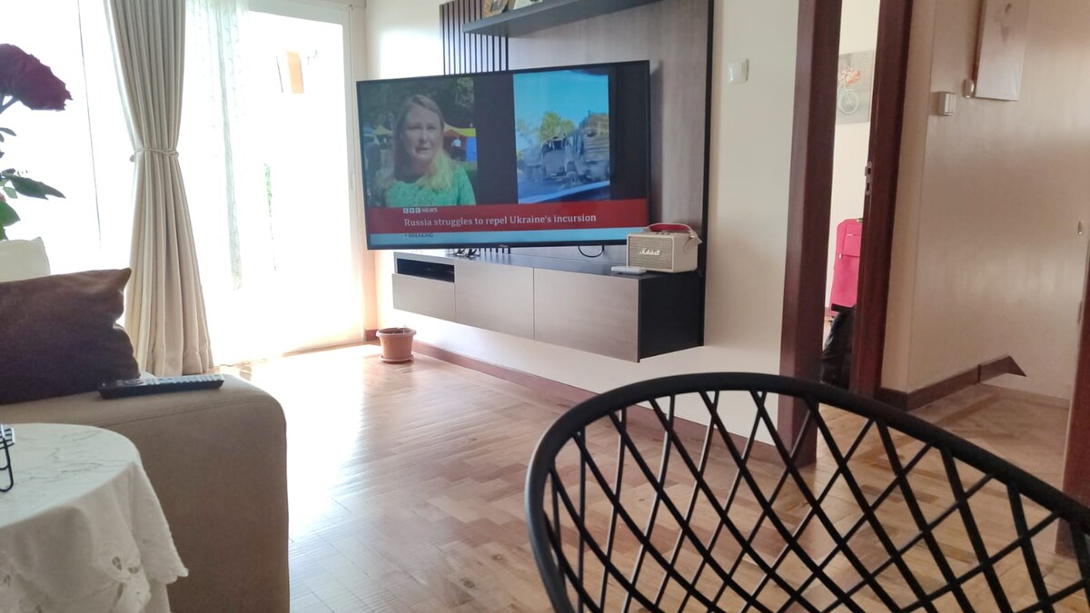 A modern living area features a wall-mounted television displaying vibrant images. Large windows allow natural light to fill the space, complemented by light-colored curtains. A cozy seating arrangement with a patterned chair and a small table enhances the room's comfort.