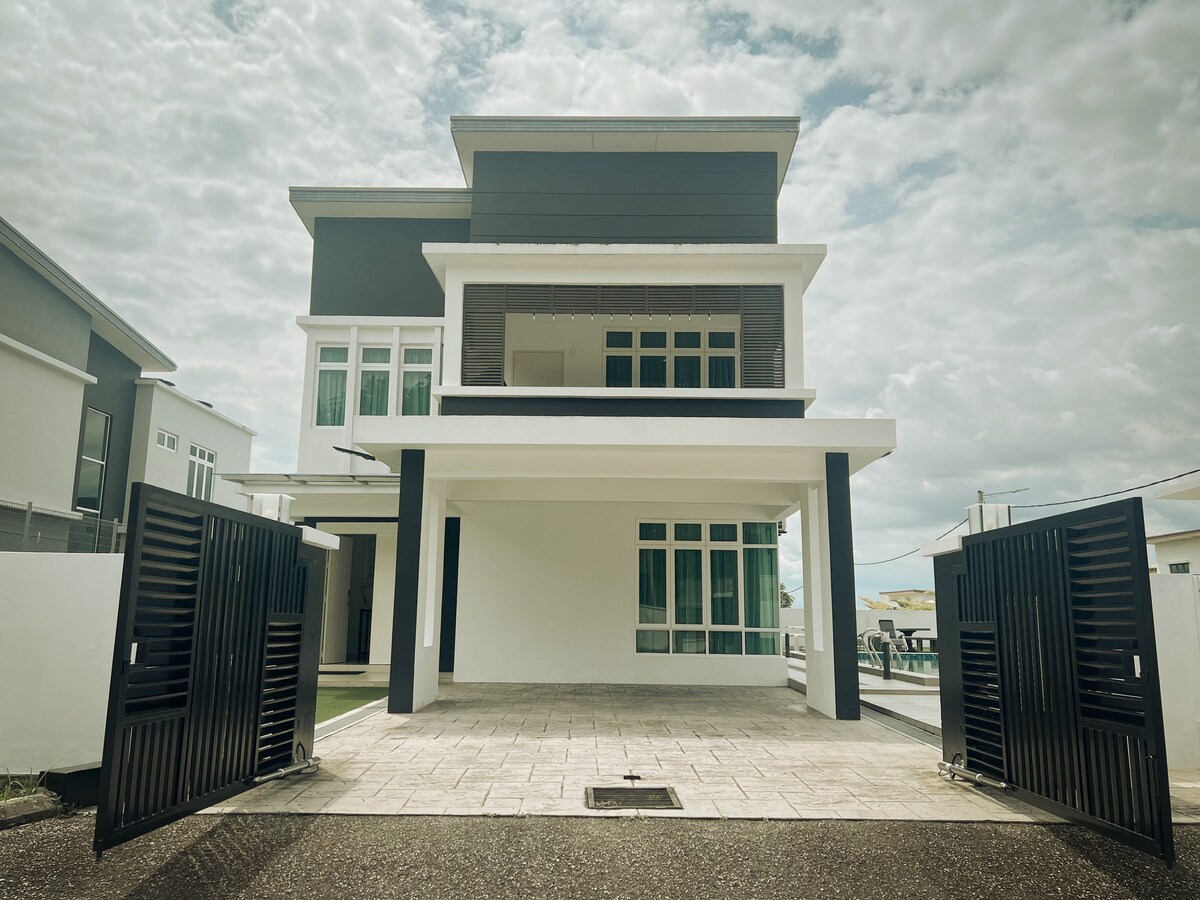 A modern two-story house is displayed with an open front yard, featuring a paved pathway leading to a large entrance. Large windows allow natural light to enter the space, while the neutral color palette adds a contemporary feel. A double-gate provides access to the property.