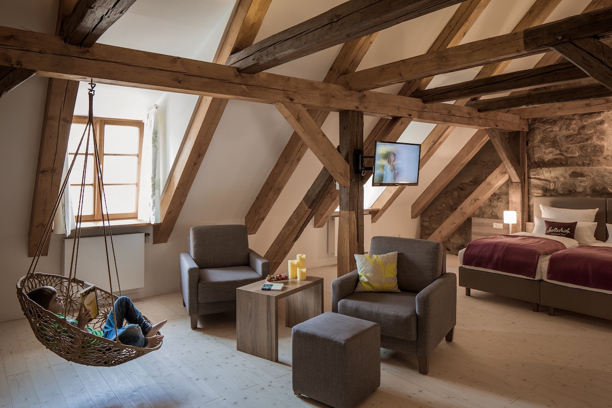 A spacious attic room features exposed wooden beams and a cozy seating area with two armchairs and a side table. A hanging chair adds a playful element, while a queen-sized bed with plush linens is positioned against a stone wall. Natural light filters through a window.