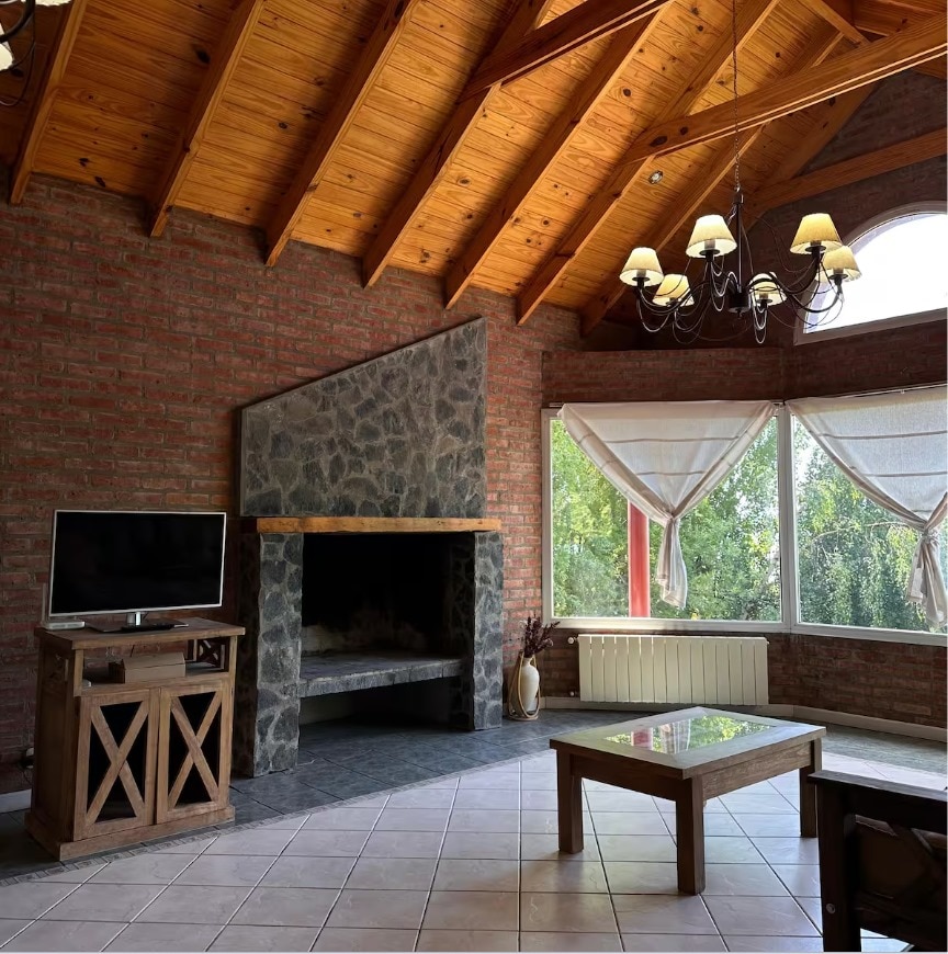 A living area features high wooden ceilings with exposed beams. A stone fireplace occupies one corner, and a television is positioned on a wooden stand. Large windows are adorned with light curtains, allowing natural light to illuminate the space.