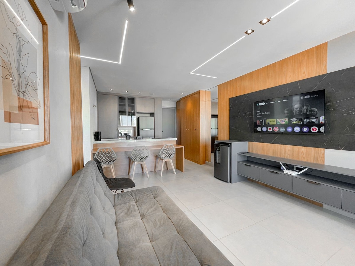 A modern living area features a sleek gray sofa and a wall-mounted television. An open kitchen with high chairs and a breakfast bar is visible, complemented by warm wooden accents. Subtle lighting highlights the space, creating a stylish and functional environment.