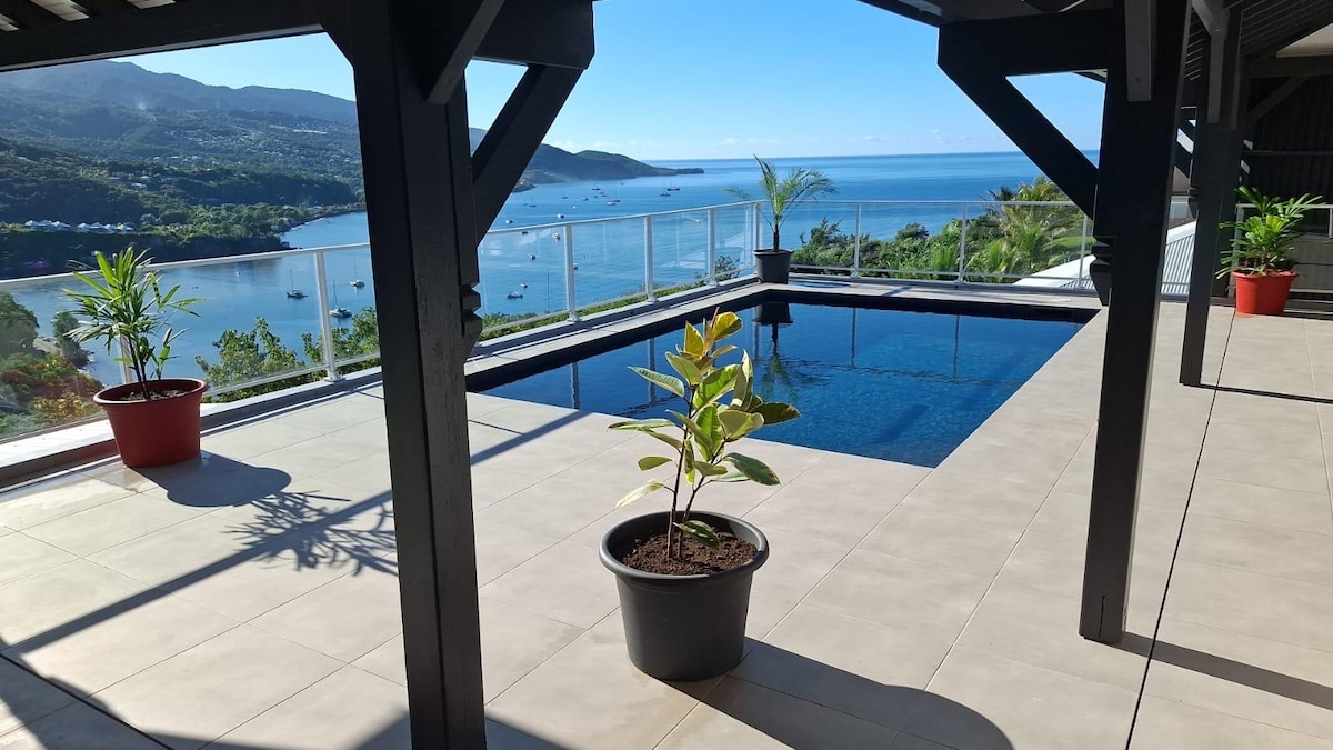 A terrace is shown featuring a swimming pool surrounded by sleek tiled flooring. Potted plants are placed strategically, and the stunning view of Malendure Bay is visible in the background, with boats gently floating on the water under a clear blue sky.