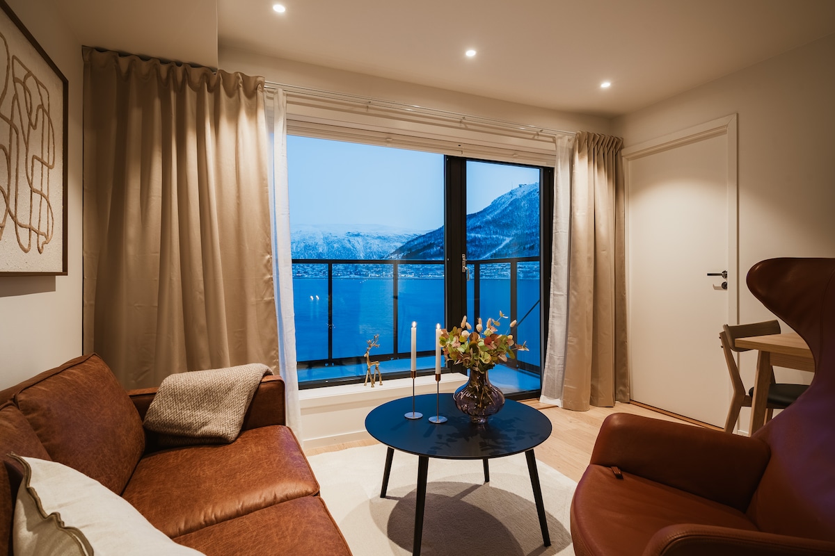A warm living room is highlighted by a large window offering views of the sea and mountains. The room features a brown leather sofa, a circular coffee table with a floral arrangement, and a comfortable chair. Soft, neutral curtains frame the window.