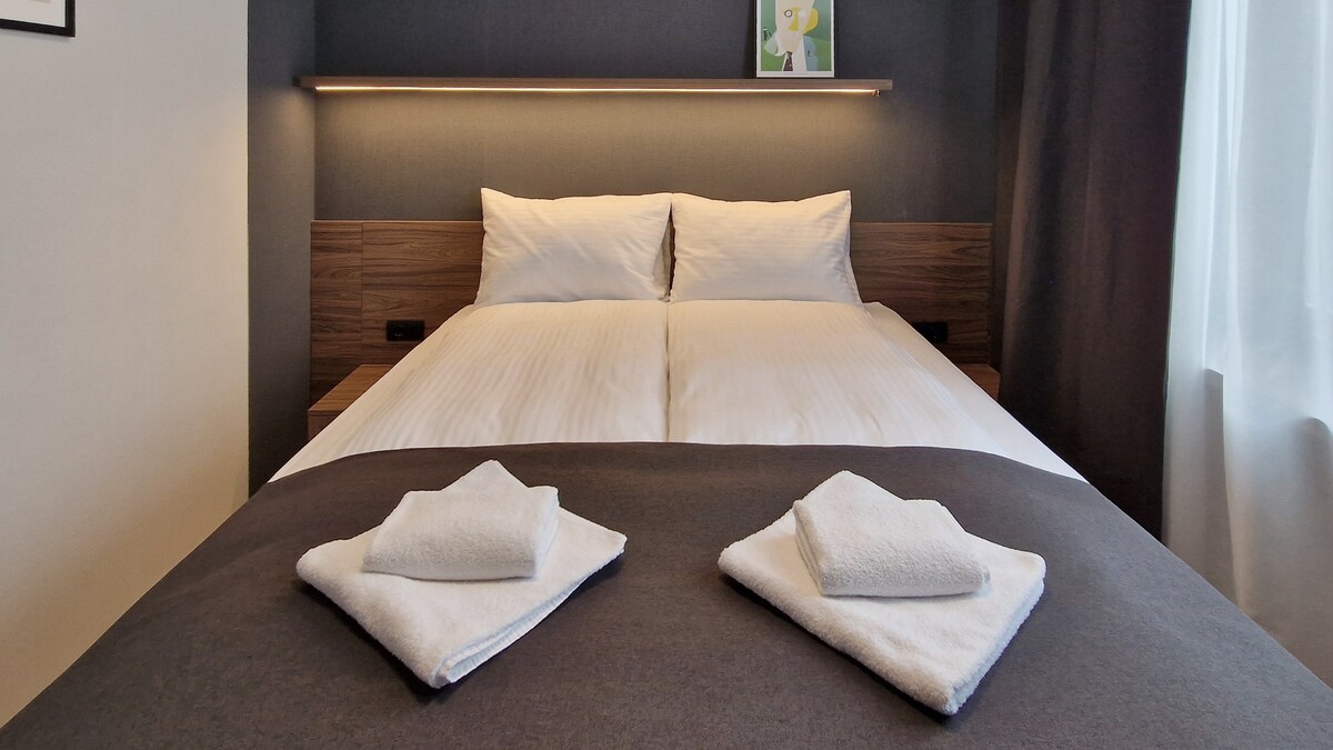 A well-appointed sleeping area features a spacious bed with crisp linens and two neatly folded towels placed on a dark bedspread. Soft lighting is provided by a wall-mounted fixture above the bed, complemented by light streaming in from a nearby window dressed with curtains.