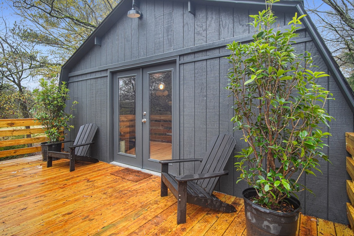 A private balcony is showcased with two dark wooden chairs positioned for relaxation. Lush potted plants frame the entrance, and a door leads back into the loft. A rich wooden deck adds a warm touch, while trees provide a natural backdrop.