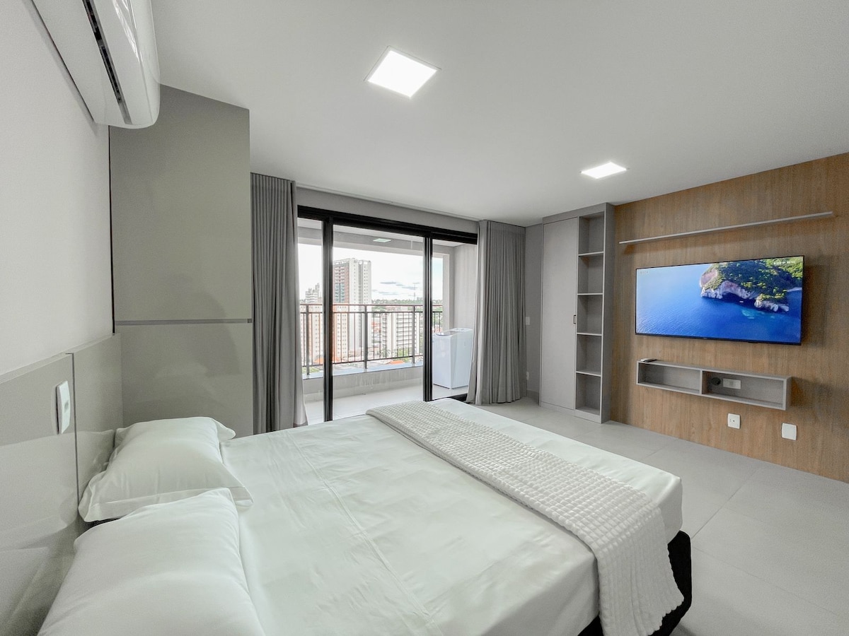 A modern bedroom features a spacious bed with crisp white linens, flanked by soft grey walls. Natural light enters through large glass doors leading to a balcony, offering views of the city. A flat-screen TV is mounted on a wooden accent wall, adding functionality to the space.