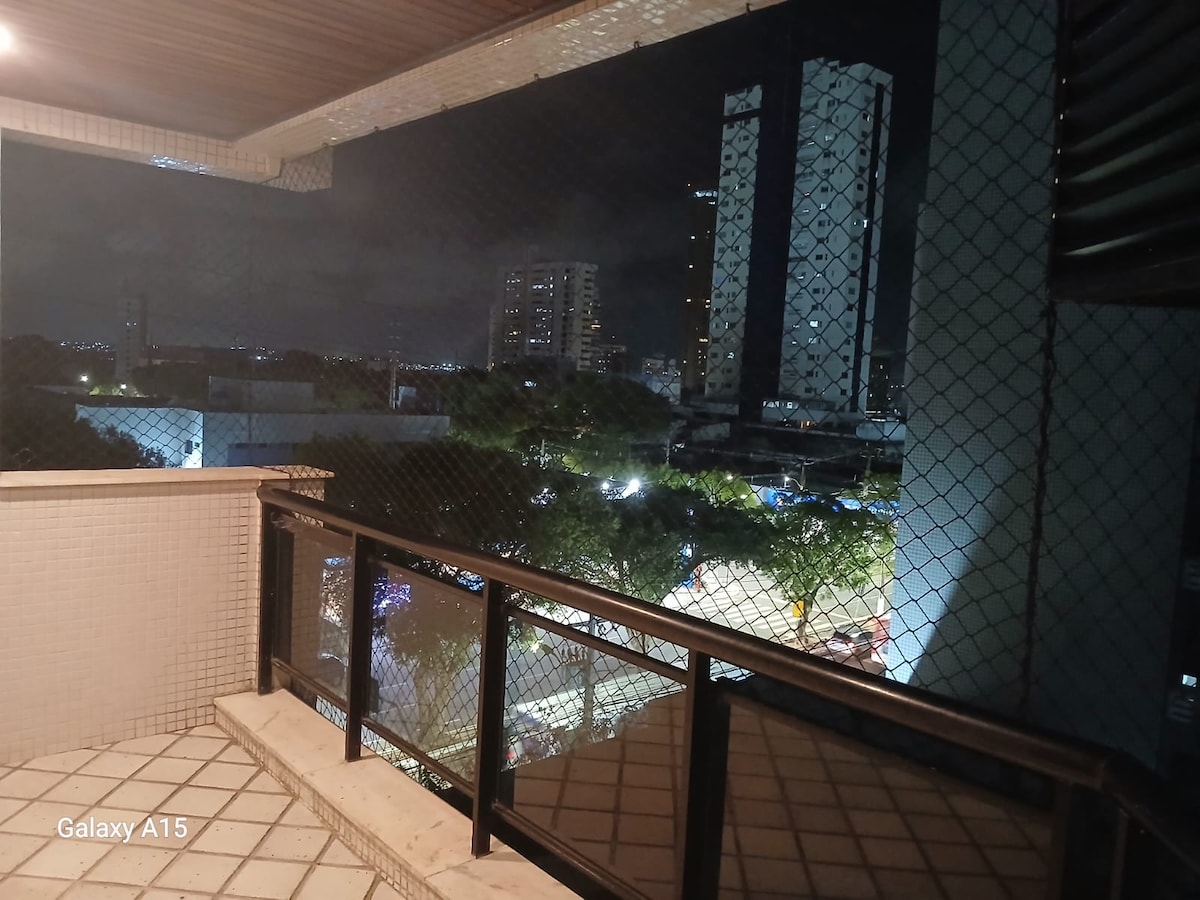 A balcony view showcases an urban landscape at night, with city lights illuminating the streets below. The railing is constructed of dark material, and a nearby window is partially visible. Surrounding buildings and greenery can be seen in the background.
