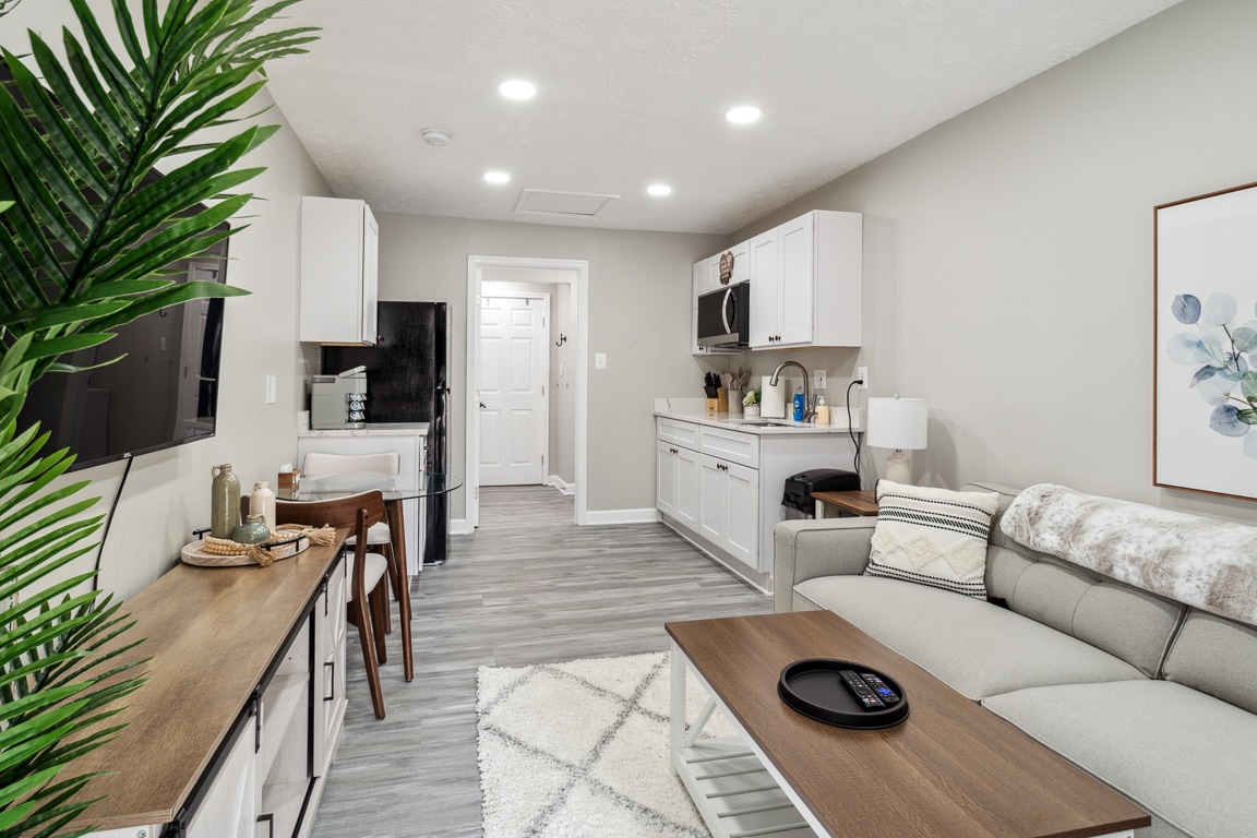 A modern living space is presented, featuring a neutral color palette and sleek design. A comfortable sectional sofa is positioned beside a coffee table, with a television mounted on the wall. The kitchenette, equipped with essential appliances, is visible in the background.