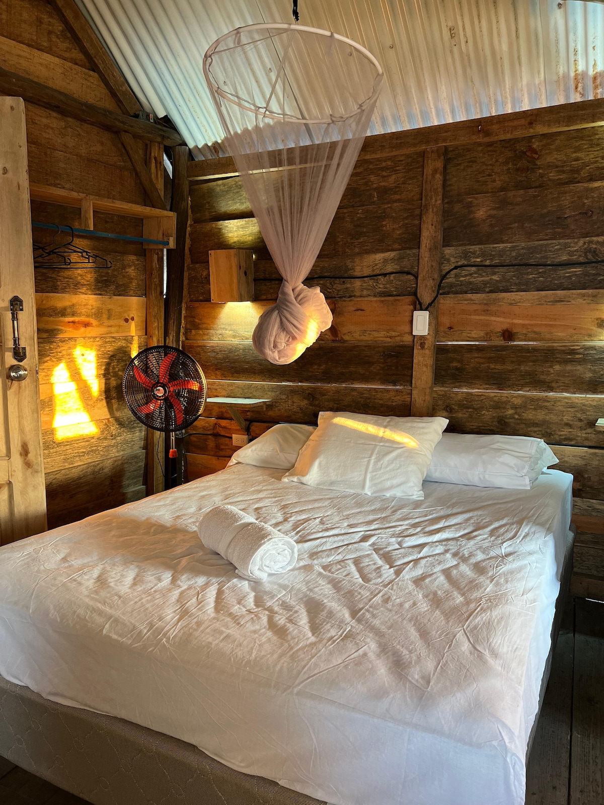 A private room is filled with rustic charm, featuring a spacious bed with white linens and a rolled towel. A fan hangs on the wall, and soft light enters through the wooden walls, creating a serene atmosphere.
