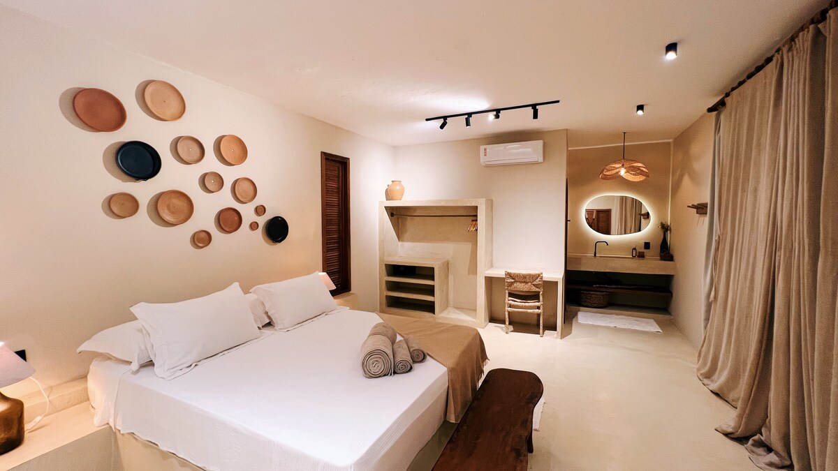 A spacious sleeping area features a queen size bed with white linens and decorative towels. The wall is adorned with various circular plates, and a warm light fixture is suspended above. A cozy seating area is provided by a wooden bench near a neutral-colored fireplace.