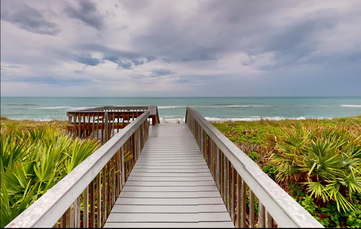 A wooden boardwalk extends through lush greenery towards the beach, leading to the calm layers of the Atlantic Ocean under a cloudy sky. Slight shades of blue and green are visible in the water, while the sandy shore meets the gentle waves in the distance.