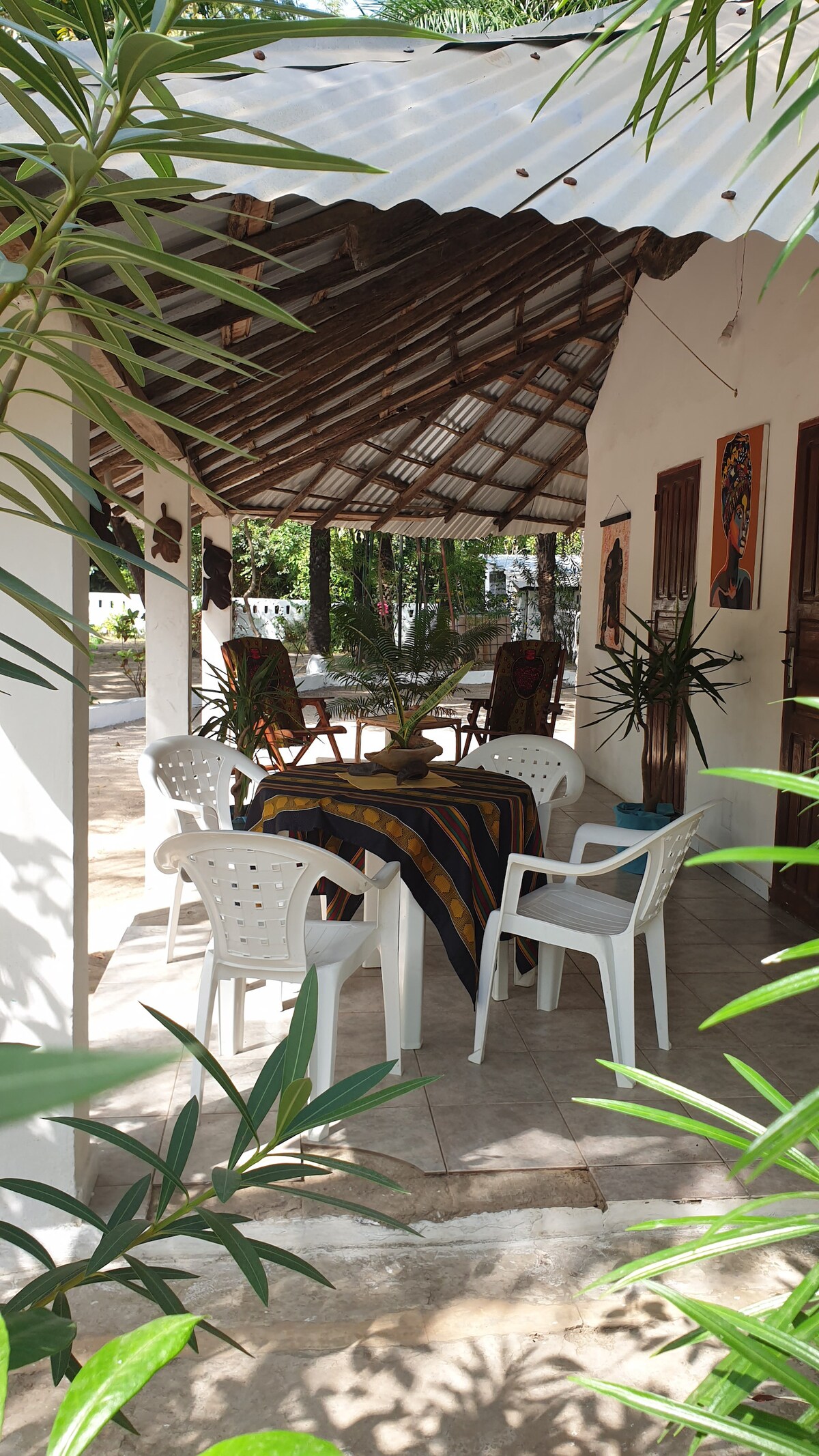 A shaded terrace area features a table draped with a colorful cloth and surrounded by multiple white chairs. Lush green plants frame the scene, creating a serene space that invites relaxation and outdoor dining.