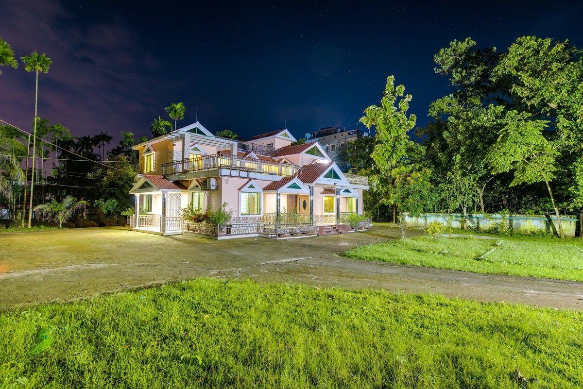 The exterior of the holiday home is illuminated at night, showcasing its inviting architecture and landscaped surroundings. A spacious yard is visible, bordered by trees, while warm lights accentuate the various entries and windows of the multi-level building.