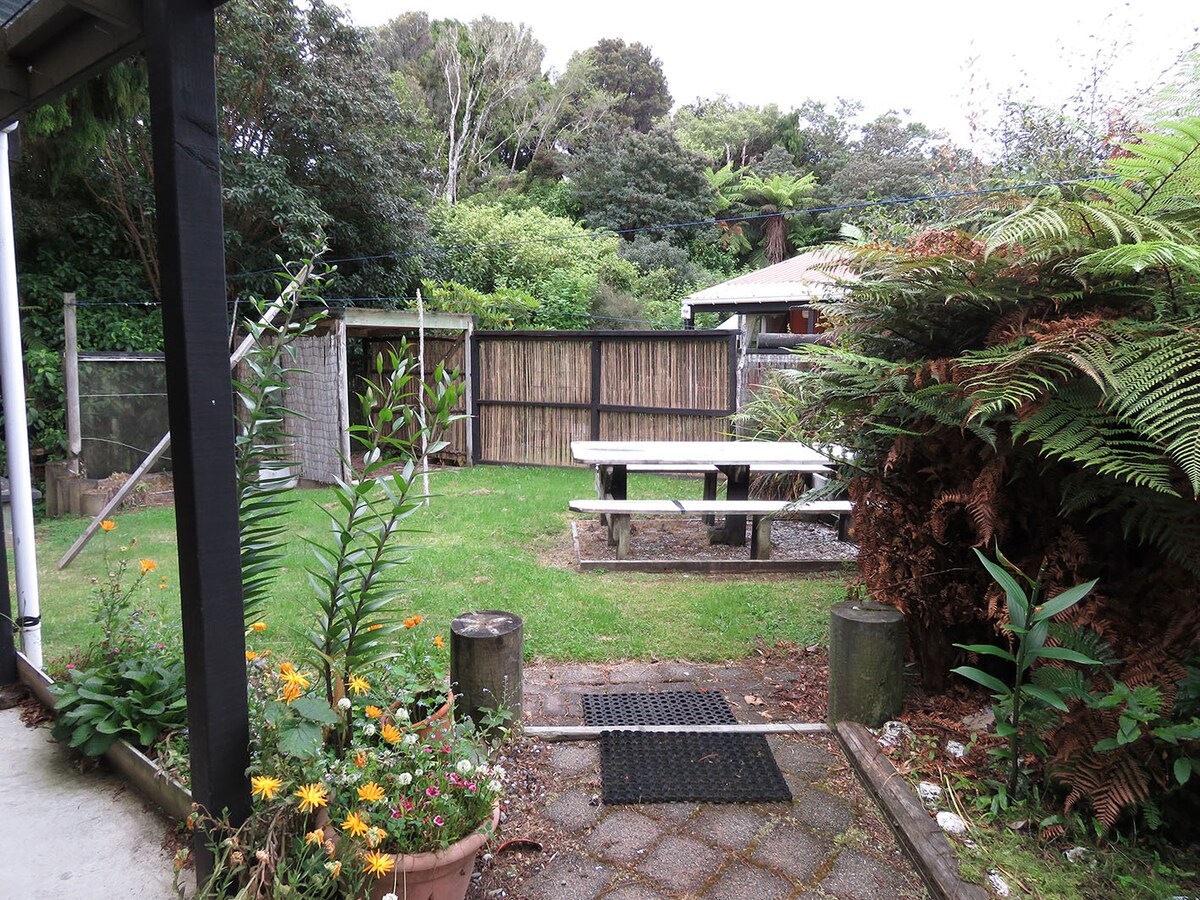A green lawn is surrounded by lush foliage, featuring a wooden picnic table positioned on a gravel area. Flowering plants add color in the foreground, while a wooden fence and trees create a natural backdrop.