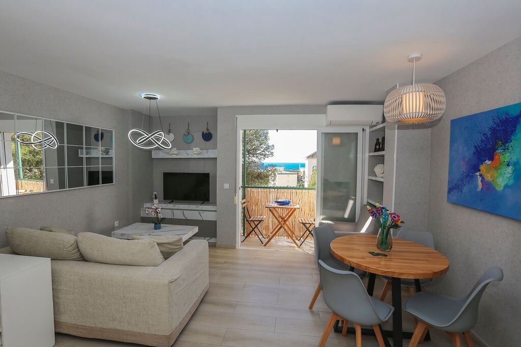 An inviting living space is highlighted by a comfortable sofa and a light wood dining table surrounded by gray chairs. A flat-screen television is mounted on the wall, while a sliding glass door offers views of the ocean and access to a small outdoor area.