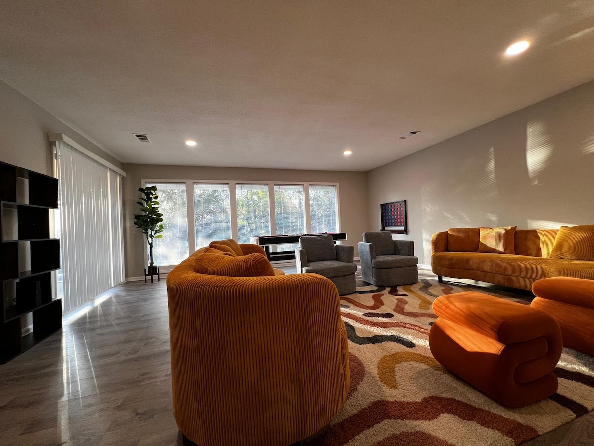 A spacious living area is filled with curved orange sofas and accent chairs, inviting relaxation. Large windows allow natural light to fill the room, while a vibrant area rug adds color to the neutral décor. A game table is positioned near the back for added entertainment.