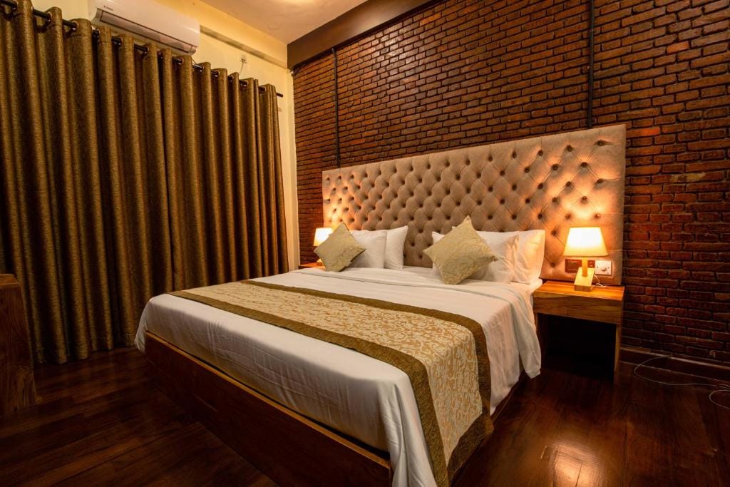 A spacious sleeping area features a plush, tufted headboard, with decorative pillows placed on the bed. Warm lighting is provided by two bedside lamps, and rich, dark curtains frame a window. Exposed brick walls add texture to the inviting environment.
