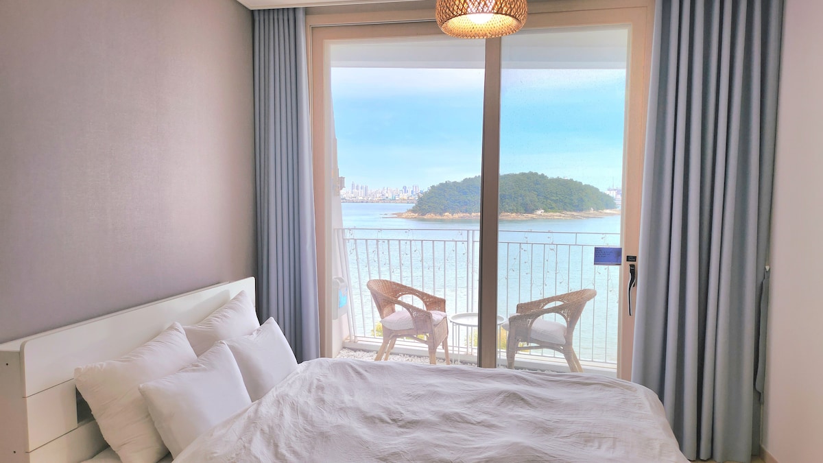 A serene bedroom features a spacious bed topped with soft white linens. Large glass doors lead to a balcony with two wicker chairs, offering a view of the tranquil ocean and a distant island, framed by light curtains that softly diffuse natural light.