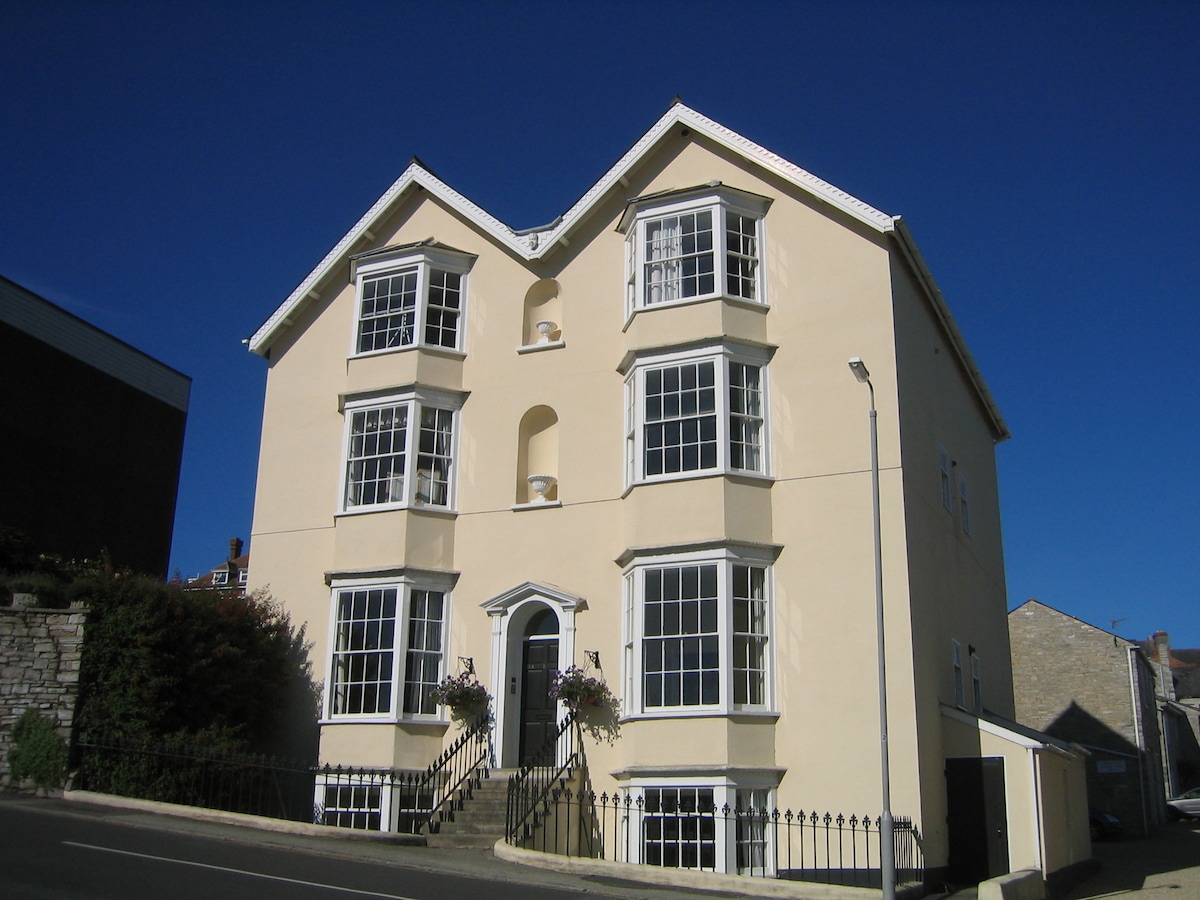 The exterior of Osborne House is displayed, featuring a symmetrical façade with large bay windows. The light-colored structure contrasts against a clear blue sky. A small set of steps leads to the front entrance, framed by decorative planters.