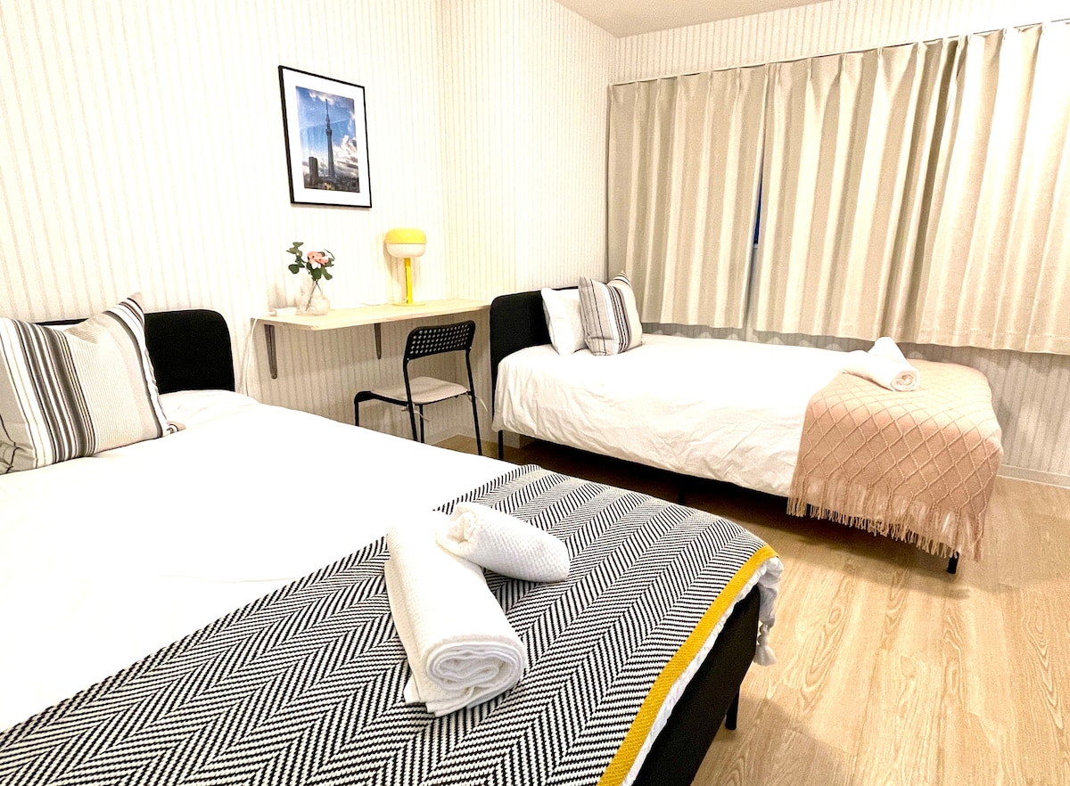 A well-lit room features two spacious beds, each adorned with fresh linens and towels. A simple desk and chair provide a cozy workspace, complemented by a decorative lamp. Soft curtains frame a window, allowing natural light to brighten the space.