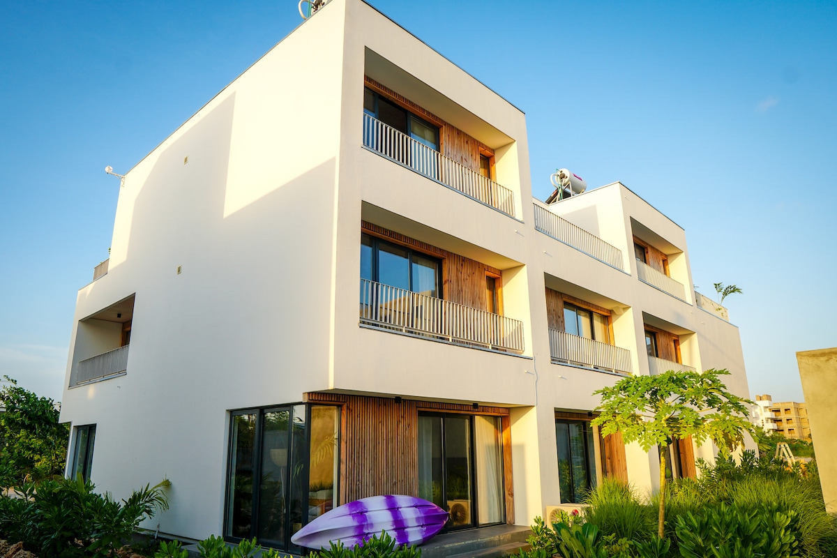 A modern timber house is portrayed, featuring multiple balconies with wooden railings. Large glass windows reflect the sunlight, and lush green landscaping surrounds the property. A surfboard is positioned in the foreground, emphasizing the home's connection to outdoor activities.