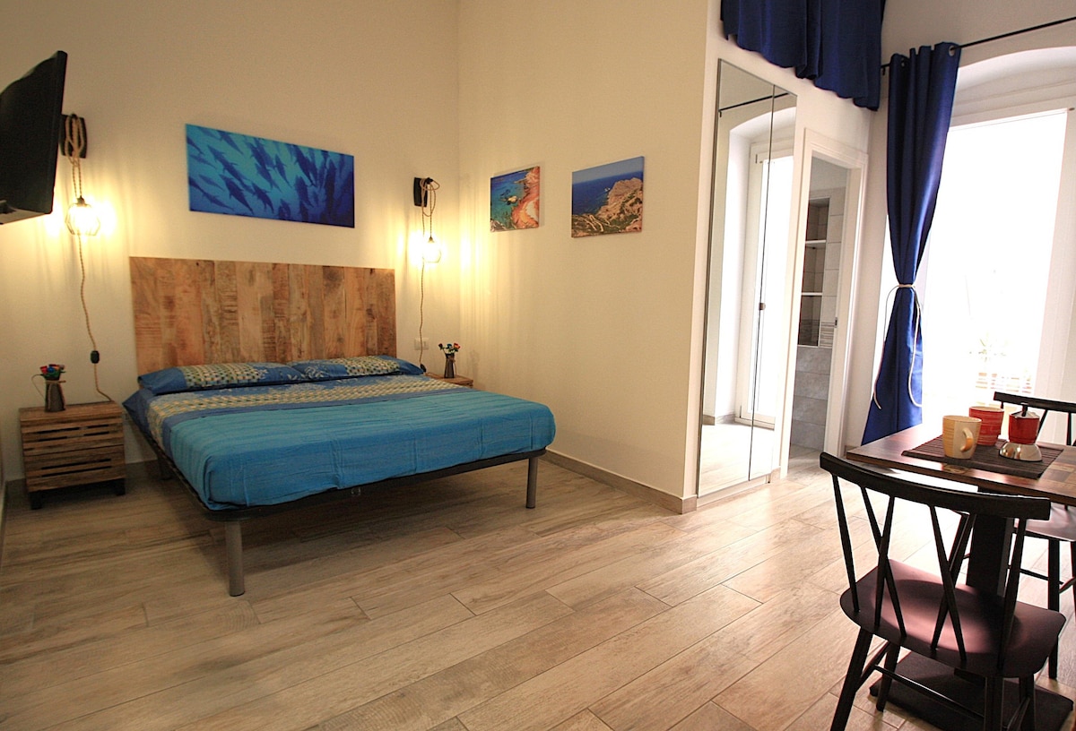 A bright sleeping area features a spacious bed with a wooden headboard, dressed in blue linens. Colorful wall art decorates the walls, while a small dining table and chairs are positioned nearby. Natural light enters through the balcony doors, complemented by dark curtains.