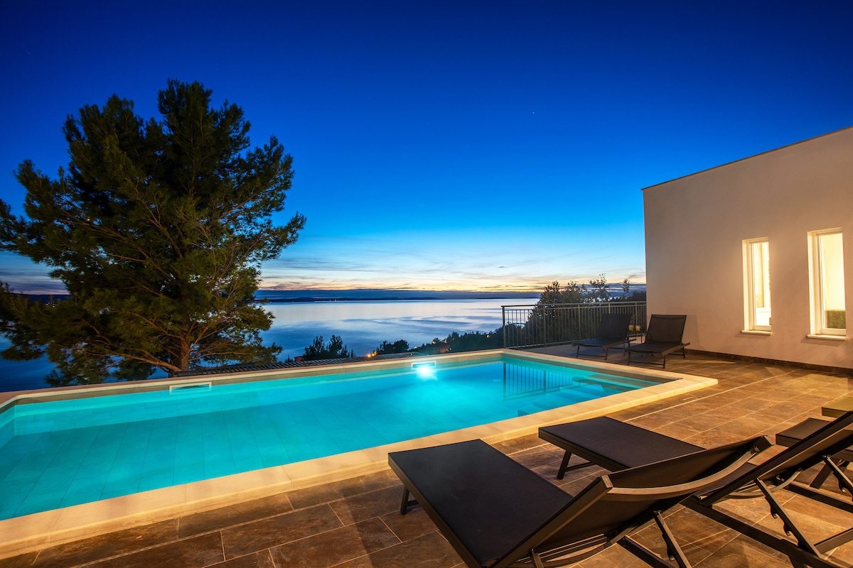 An outdoor area features a private pool with clear blue water, surrounded by sleek loungers. The tranquil sea is visible in the background under a deep blue sky transitioning to twilight, framed by lush trees providing a serene atmosphere.