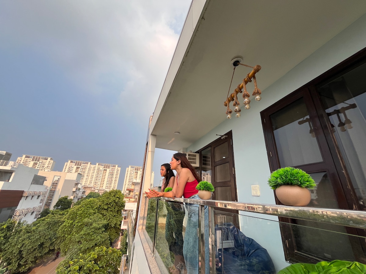 A balcony offers a view of the surrounding cityscape, with greenery visible below. A decorative light fixture hangs from the ceiling, while potted plants are placed on the glass railing. The sky displays a blend of clouds and sunlight.