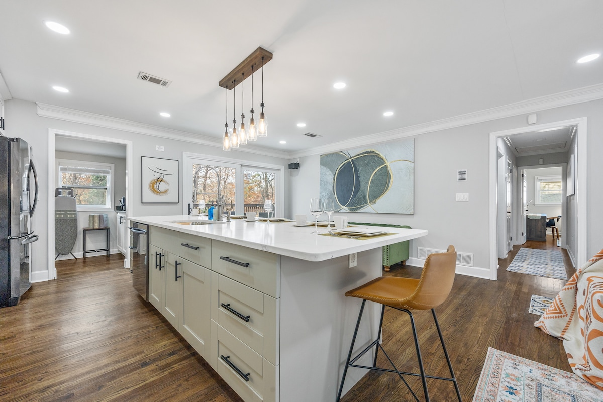 An open concept kitchen showcases a spacious island with bar seating and contemporary light fixtures overhead. Natural light fills the room through large windows, highlighting the modern color palette and design elements, including artwork on the walls and sleek kitchen appliances.
