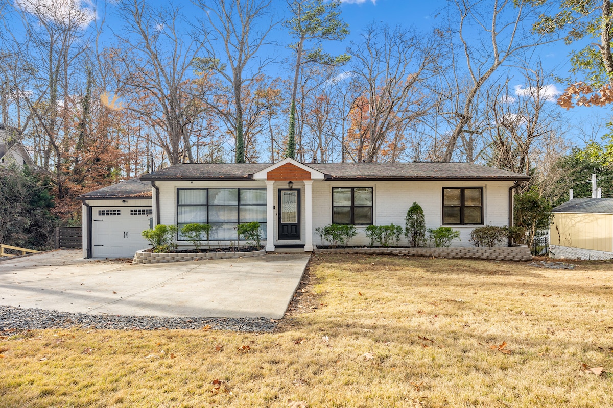 The single-story home is designed with a classic façade, featuring a central entrance framed by a decorative overhang. A well-maintained front yard with shrubs and a gravel driveway leads to an attached garage. Surrounding trees enhance the serenity of the environment.
