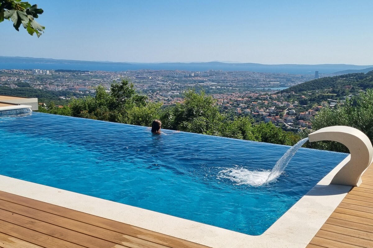 A private infinity pool overlooks stunning panoramic views of the town below and the Adriatic Sea in the distance. Lush greenery surrounds the pool area, providing a serene environment. Water flows gently from an elegant spout into the sparkling blue water.