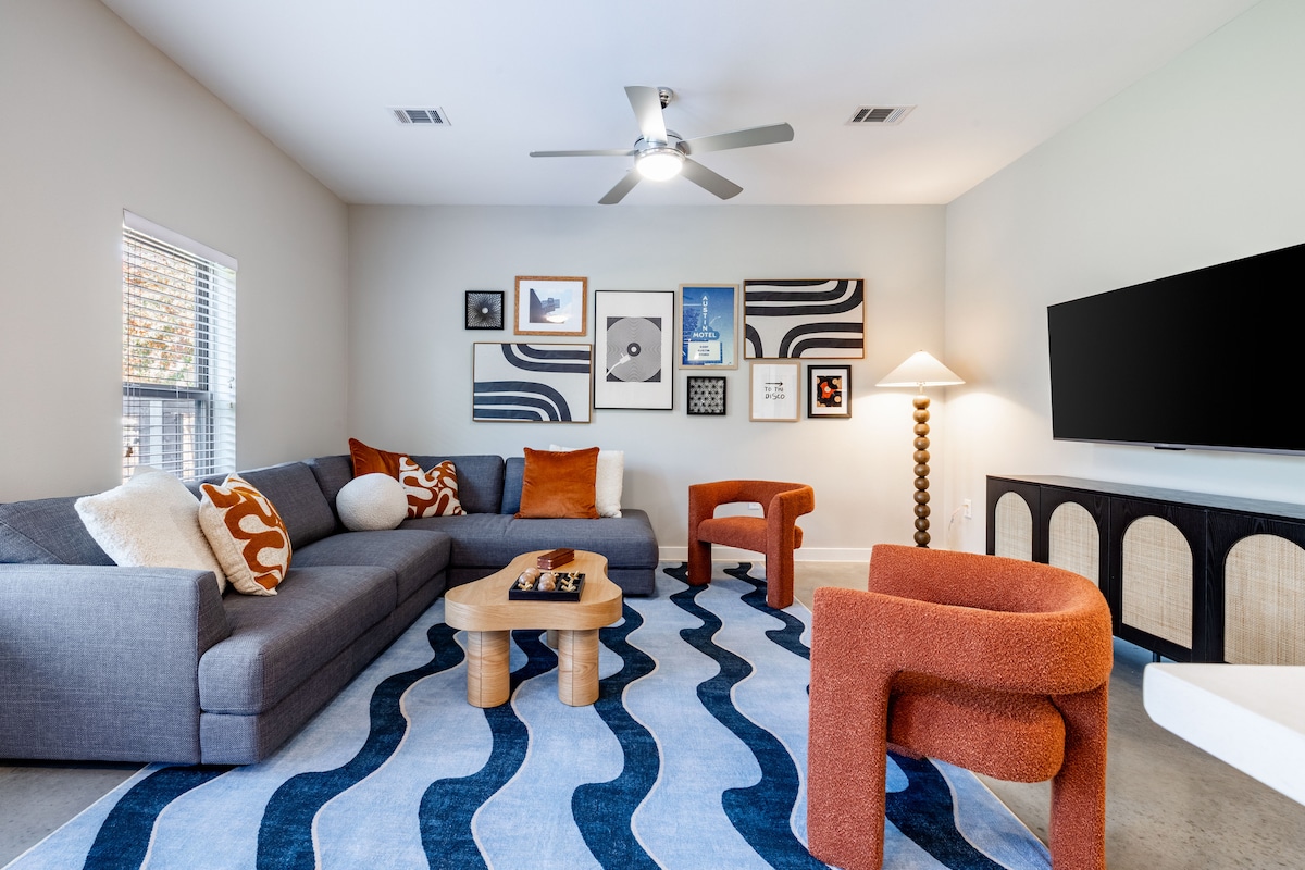 A modern living area features a sectional sofa with decorative pillows, complemented by two orange accent chairs. Art pieces are arranged on the light-colored walls, and a large area rug with a wave pattern anchors the space. A flat-screen TV is mounted on the wall.